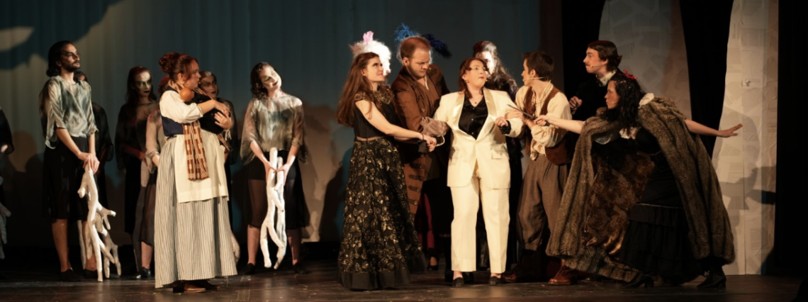 Cast members of "Into the Woods" including members of the ensemble who are holding decorative tree branches, Karleigh Martin as The Baker’s Wife who is holding a doll, Aoibhinn Macken-Luyt as The Witch, Jack French as Cinderella’s Father, Taylor Pike as the Narrator and Charly Graham as Little Red Riding Hood, on stage at War Memorial Hall at the University of Guelph. The stage is set as a forest with trees. Photo credit: Joshua Lonuzzo 
